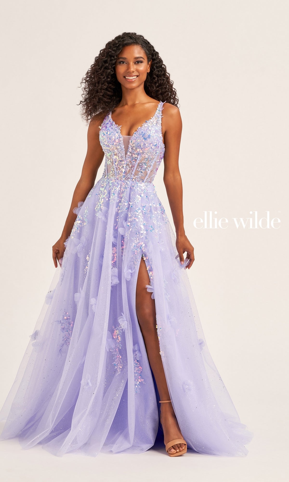 A woman models the Long Glitter Prom Ball Gown EW35047 by Ellie Wilde—a lavender, floral-embellished gown with a sheer bodice and high slit—standing against a plain background. The